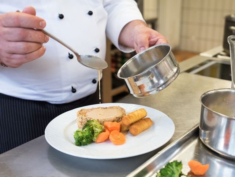 Nahaufnahme: ein Koch gibt Soße auf einen Teller mit Fleisch, Kroketten und Gemüse
