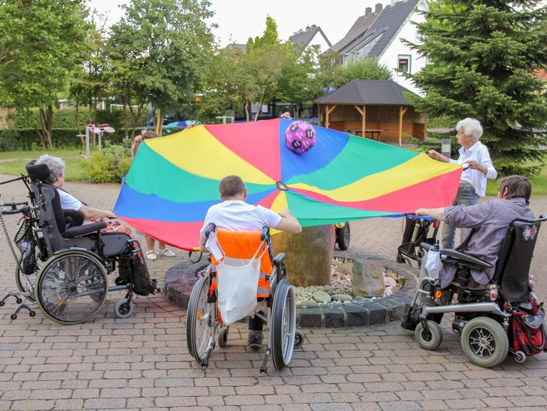 Männer und Frauen teils im Rollstuhl spielen in der Auffahrt mit einem großen bunten Tuch und einem Ball darin