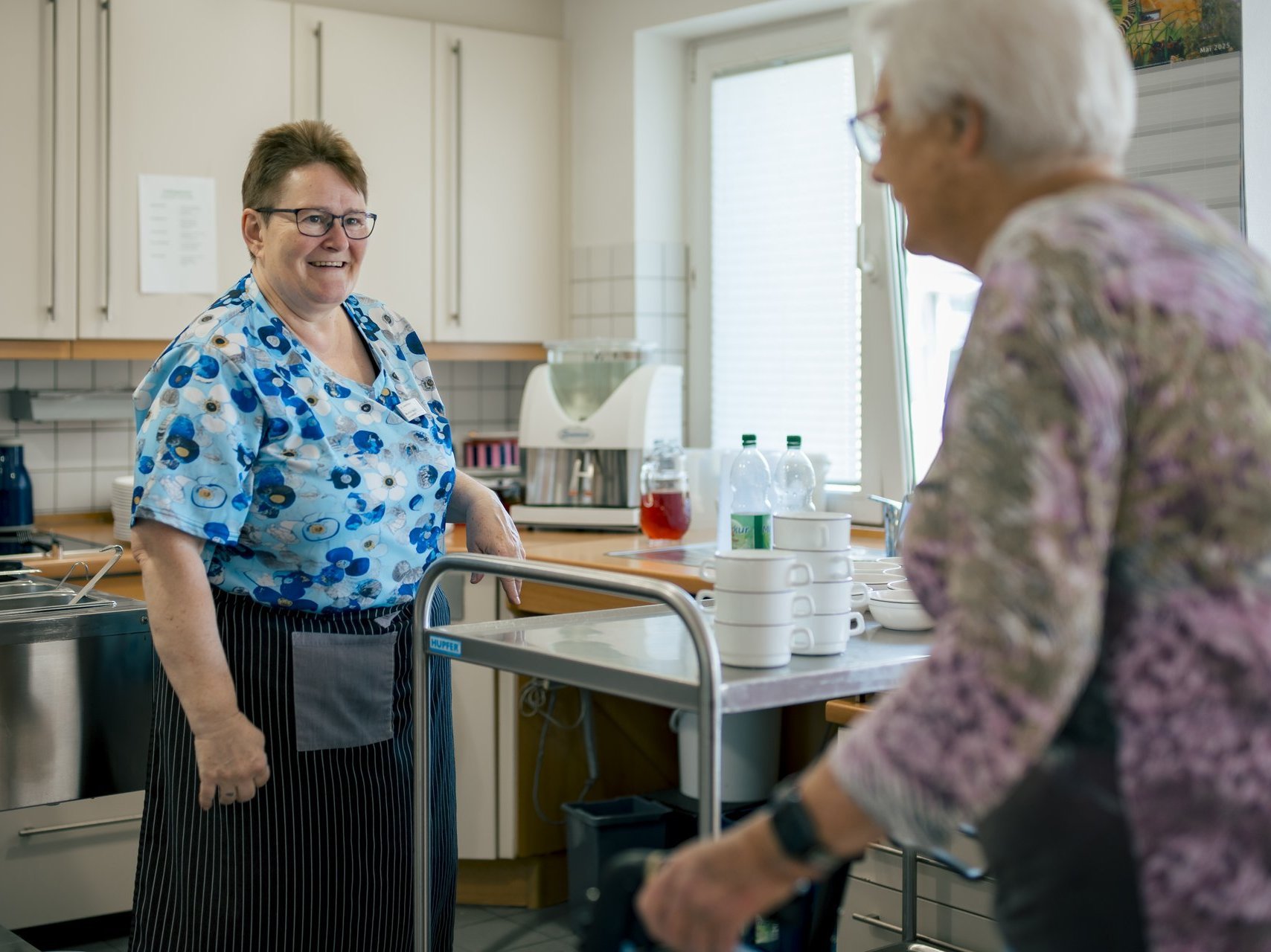Eine Mitarbeiterin der Hauswirtschaft unterhält sich lachend mit einer Seniorin.