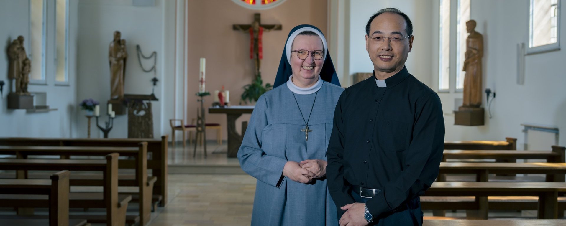 Der Hausgeistliche und eine Ordensschwester stehen in der Hauskapelle und lächeln in die Kamera.