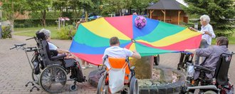 Männer und Frauen teils im Rollstuhl spielen in der Auffahrt mit einem großen bunten Tuch und einem Ball darin