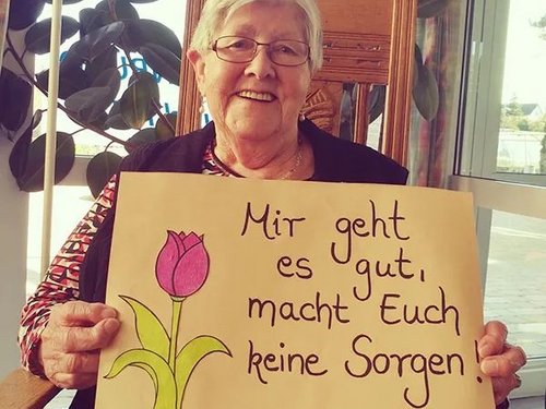 eine Frau hält ein ein Plakat mit einer Blume und der Aufschrift: "Mir geht es gut, macht Euch keine Sorgen!"