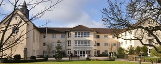 Über eine Rasenfläche blickt man auf ein U-Förmiges Gebäude in Beige. Auf dem Dach des linken U-Schenkels befindet sich ein Türmchen mit Kreuz. Mittig angeordnet ist der Eingangsbereich über dem der Schriftzug "St. Josefshaus" zu lesen ist.