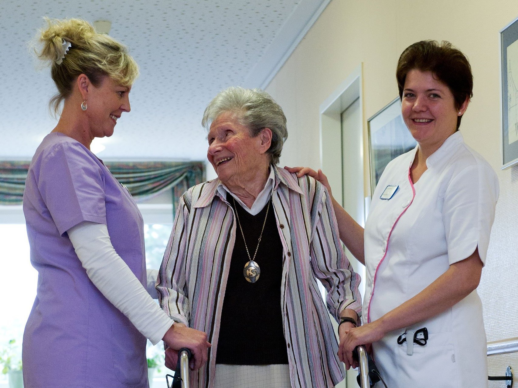 Zwei Pflegerinnen helfen einer alten Frau mit Gehhilfe