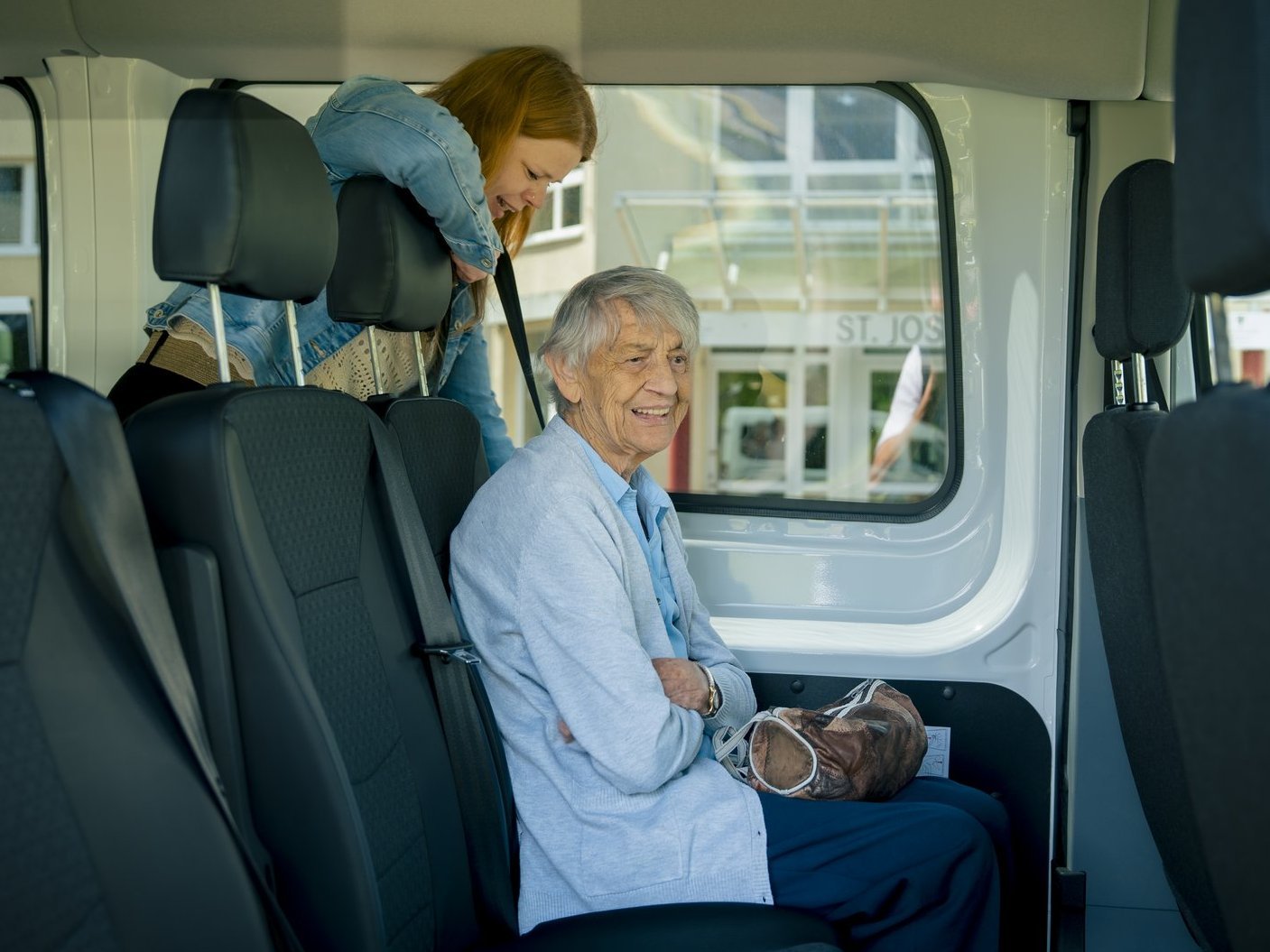 Eine Seniorin sitzt im Fahrzeug der Altenhilfe und lacht in die Kamera. Eine Mitarbeiterin schnallt sie an.