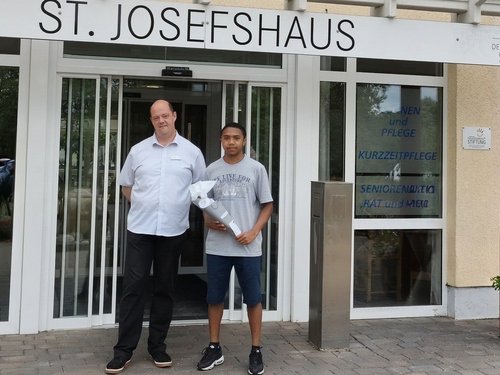 Zwei Männer stehen vor dem Haupteingang des St. JosefsHaus. Der Mann rechts hält ein gerolltes Dokument. Über der Glastür ist der Schriftzug „ST. JOSEFSHAUS“ deutlich sichtbar.