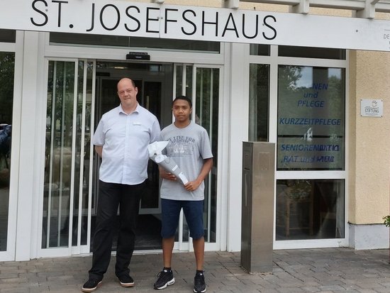 Zwei Männer stehen vor dem Haupteingang des St. JosefsHaus. Der Mann rechts hält ein gerolltes Dokument. Über der Glastür ist der Schriftzug „ST. JOSEFSHAUS“ deutlich sichtbar.