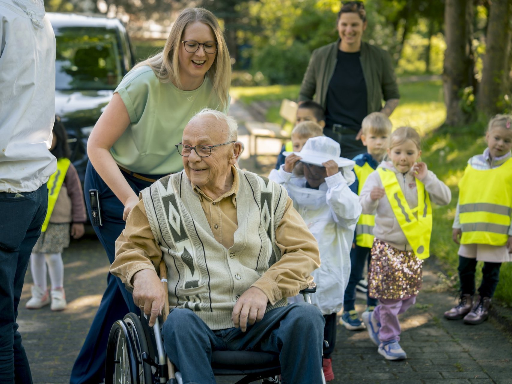 Eine Mitarbeiterin und ein Senior im Rollstuhl befinden sich in einem Park. Sie sind umgeben von Kindern und anderen Menschen.