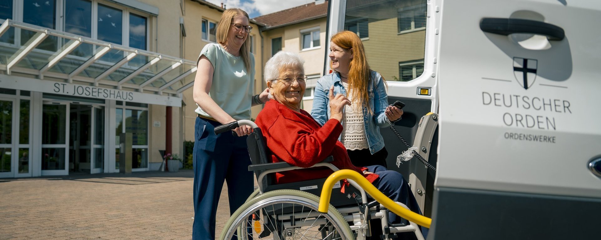 Eine im Rollstuhl sitzende Seniorin lacht in die Kamera, während zwei Mitarbeiterinnen ihr dabei helfen, in das Fahrzeug der Altenhilfe einzusteigen.