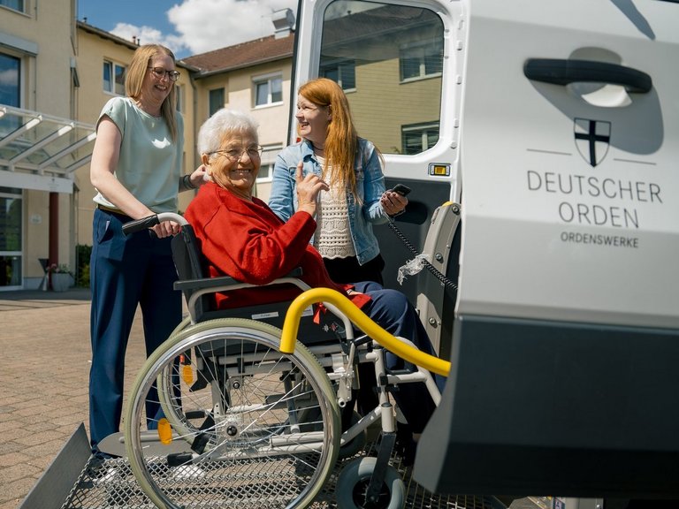 Eine im Rollstuhl sitzende Seniorin lacht in die Kamera, während zwei Mitarbeiterinnen ihr dabei helfen, in das Fahrzeug der Altenhilfe einzusteigen.