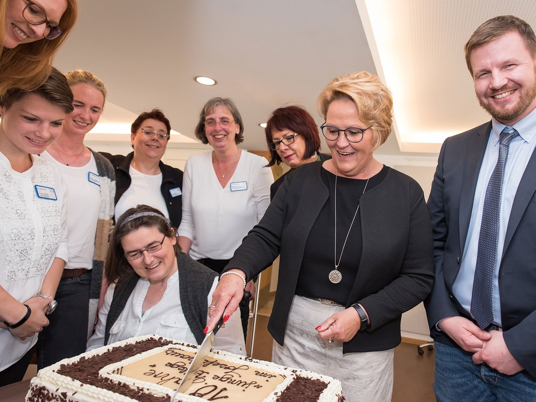 Eine Frau durchschneidet eine große Torte - um sie herum stehen viele Pflegerinnen und ein Pfleger. Auf der Torte steht etwas mit 10 Jahre...