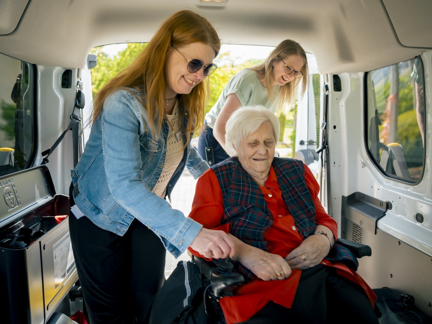 Zwei Mitarbeiterinnen unterstützen eine Seniorin im Rollstuhl, sich im Fahrzeug der Altenhilfe anzuschnallen.