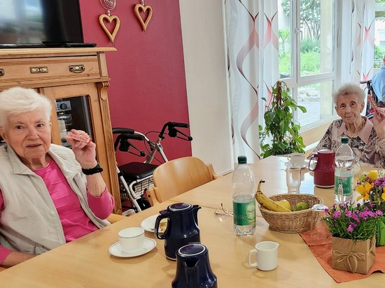 Zwei ältere Damen sitzen an einem hellen Holztisch. Vor ihnen stehen Kaffeetassen, eine blaue Kanne und Wasserflaschen. Ein Korb mit Bananen und eine kleine Blumenvase zieren die Tischmitte. Im Hintergrund ist ein großes Fenster sowie eine weitere Person im Rollstuhl zu sehen.