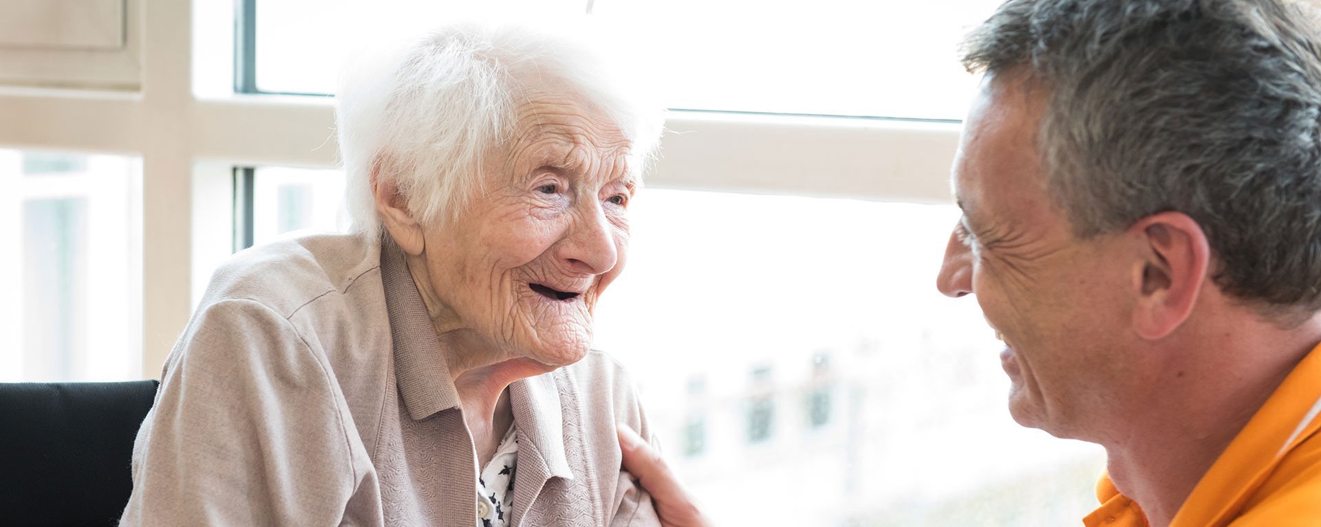 Ein Pfleger kniet vor einer alten Frau, die im Rollstuhl sitzt - beide lachen