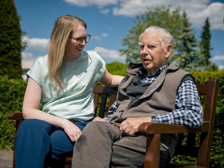 Ein Senior und eine Mitarbeiterin sitzen auf einer sonnigen Bank und unterhalten sich.