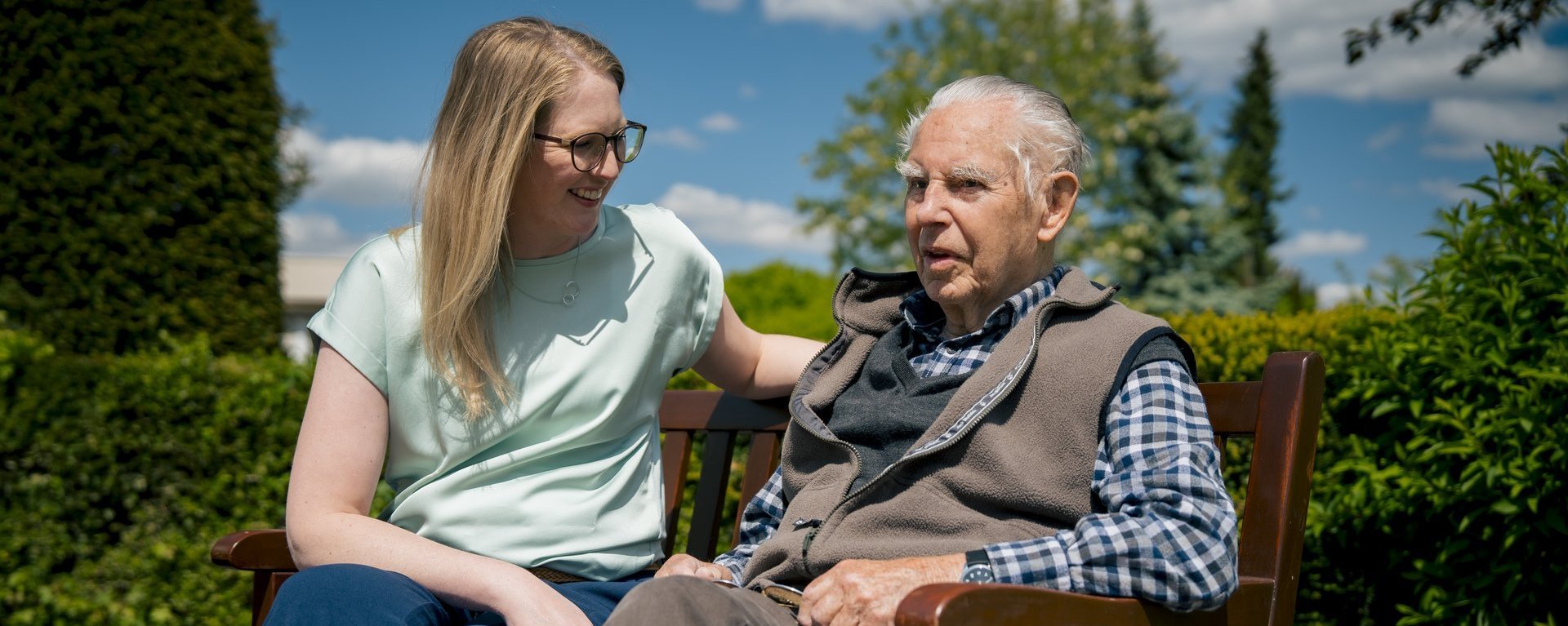 Ein Senior und eine Mitarbeiterin sitzen auf einer sonnigen Bank und unterhalten sich.