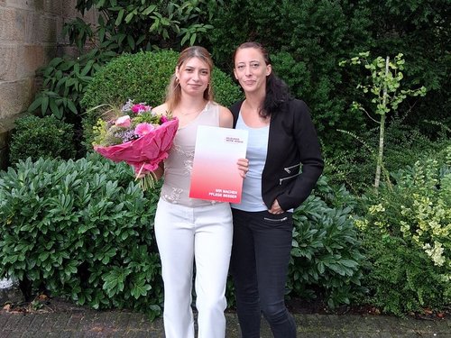 Zwei junge Frauen stehen vor einer Steinmauer und Hecken. Die linke Frau hält einen Blumenstrauß und ein Zertifikat, was auf eine Verleihung hindeutet. Beide lächeln in die Kamera.