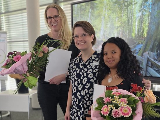 Drei lächelnde Frauen stehen nebeneinander und halten Blumensträuße. Zwei von ihnen halten zusätzlich weiße Zertifikate in den Händen. Sie befinden sich in einem hellen Innenraum, was auf eine feierliche Übergabe oder Auszeichnung hindeutet.