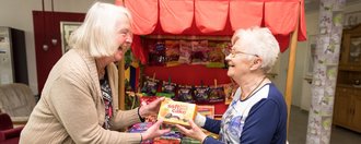 zwei ältere Frauen stehen an einem kleinen Süßigkeitenstand, lachen und halten eine Packung soft cake (orange) in den Händen