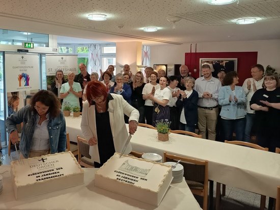 Zwei Frauen schneiden große Torten an einem Tisch, während im Hintergrund eine Gruppe von Menschen applaudiert. Die Torten gratulieren zum 20-jährigen Trägerschaftsjubiläum und zum 60-jährigen Bestehen des St. Josefshauses.