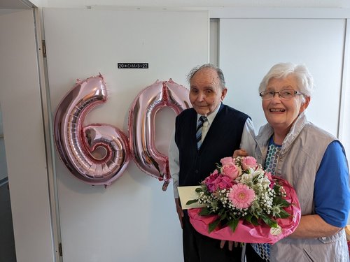 Mann und Frau vor Tür mit Luftballon "60" die Frau hält einen Blumenstrauß