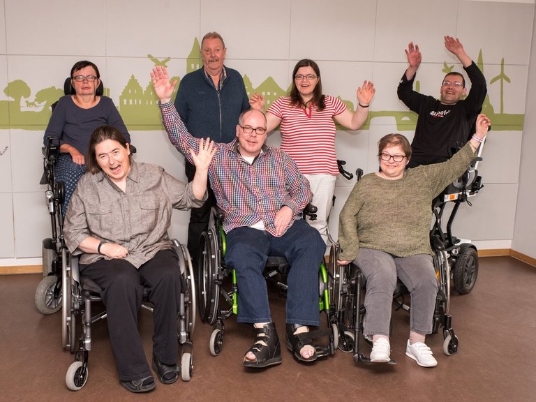 Gruppenfoto: Eine Gruppe Männer und Frauen stehen oder sitzen im Rollstuhl und winken in die Kamera