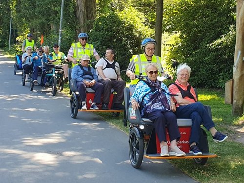 Eine Gruppe von Menschen fährt in einer Reihe von Lastenrädern auf einem asphaltierten Weg durch einen Park. Die Fahrer tragen gelbe Warnwesten, während ältere Passagiere entspannt in den vorderen Sitzen der Fahrzeuge Platz genommen haben.
