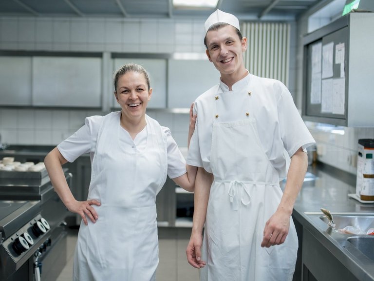 Zwei Mitarbeitende stehen in der Küche und lachen in die Kamera.