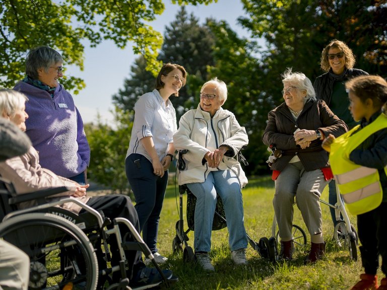 Mitarbeitende, Senioren und Kinder stehen auf einer Wiese und unterhalten sich.